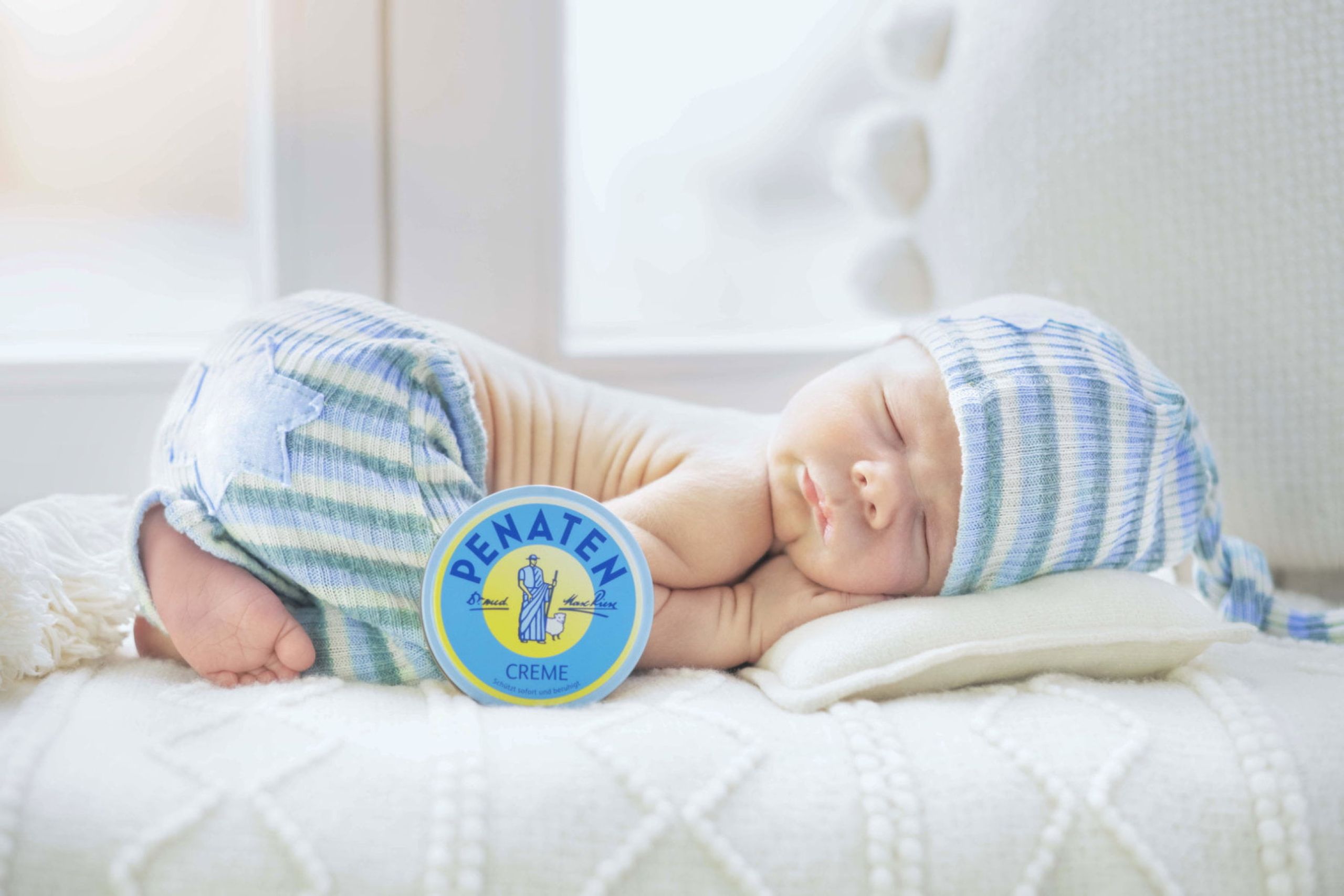 Ein Baby liegt mit einer blauen Mütze und einem blauen Höschen auf einer Decke. Davor steht eine Dose Penaten Creme. Im Hintergrund ist ein Fenster.